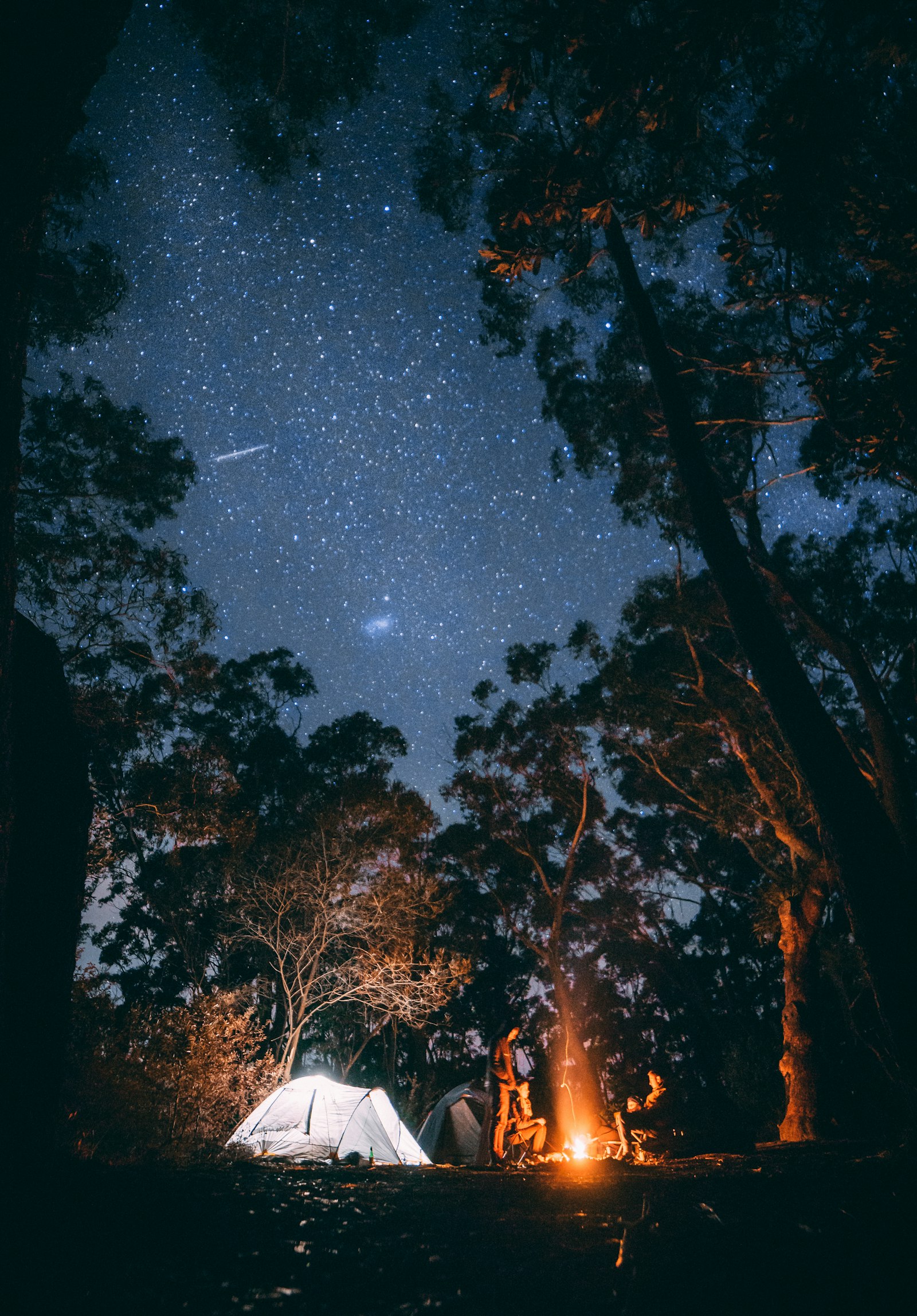 Campamento nocturno con casa de campaña iluminada y fogata bajo un cielo estrellado